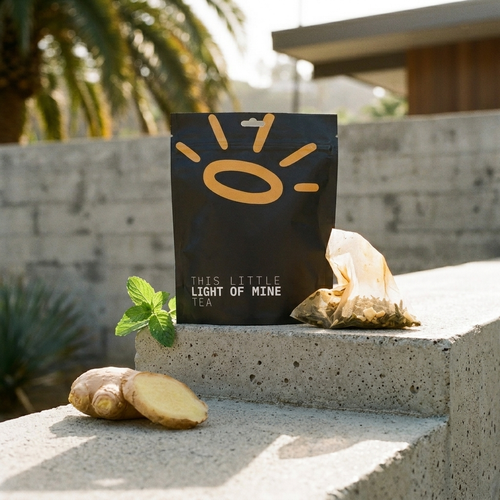 A black tea pouch labeled 'THIS LITTLE LIGHT OF MINE TEA' stands on a concrete ledge outdoors, with loose tea bags, ginger root, and mint leaves nearby. The background shows blurred palm trees and a building exterior.