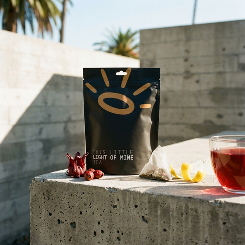 A black stand-up pouch of tea labeled 'THIS LITTLE LIGHT OF MINE TEA' stands on a concrete ledge outdoors, with a glass of red tea, tea bags, and dried red flowers nearby. The background shows blurred concrete walls and palm trees.