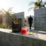 A black pouch of Chic Bottle Light of Wine Tea stands on a concrete ledge next to a clear glass mug filled with a red beverage, with a blurred person walking in the background outdoors.