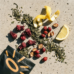 An overhead flat-lay of loose herbal tea ingredients on a textured light stone surface: dried green leaves, red hibiscus flowers, dried citrus peel, red berries, and a lemon wedge, with a black tea bag pouch shown in the lower left.