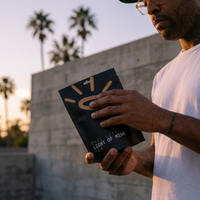 A man in a white t-shirt holds a black pouch of 'This Little Light of Mine Tea' outdoors at sunset, with palm trees and a concrete wall in the background.