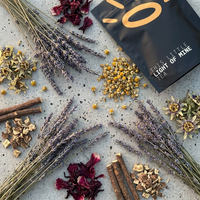 A styled flat-lay of loose-leaf tea ingredients and packaging on a light gray textured surface. A black pouch labeled 'THIS LITTLE LIGHT OF MINE' is visible, surrounded by dried lavender sprigs, chamomile flowers, cinnamon sticks, and dark red hibiscus petals.