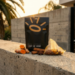 A black stand-up pouch labeled 'THE LITTLE LIGHT OF MINE TEA CO.' stands on a concrete ledge outdoors, with turmeric roots, cinnamon sticks, star anise, and a small clear bag of loose tea ingredients arranged in front.
