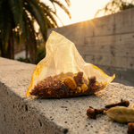 A small translucent tea bag filled with loose-leaf herbal tea and spices sits on a concrete ledge outdoors at sunset, with palm trees and a wall blurred in the background.