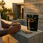 A person holding a cup of tea on a concrete balcony at sunset, with a black stand-up pouch labeled 'THE LITTLE SHOT OF MINE' and a cinnamon stick beside it.