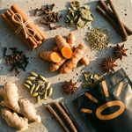 A styled flat-lay of whole spices and herbs on a speckled stone surface, including turmeric roots, cinnamon sticks tied with twine, star anise, cardamom pods, cloves, dried leaves, and a black pouch with a gold sun-like logo.