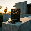 A black stand-up pouch of tea labeled 'THIS LITTLE LIGHT OF MINE TEA' stands on a concrete ledge outdoors at sunset, with a small tea bag, loose tea leaves, and two brown tea bags beside it.